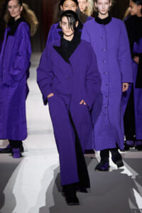 Models in matching long purple coats walk the runway, black outfits visible at the collar and cuffs.
