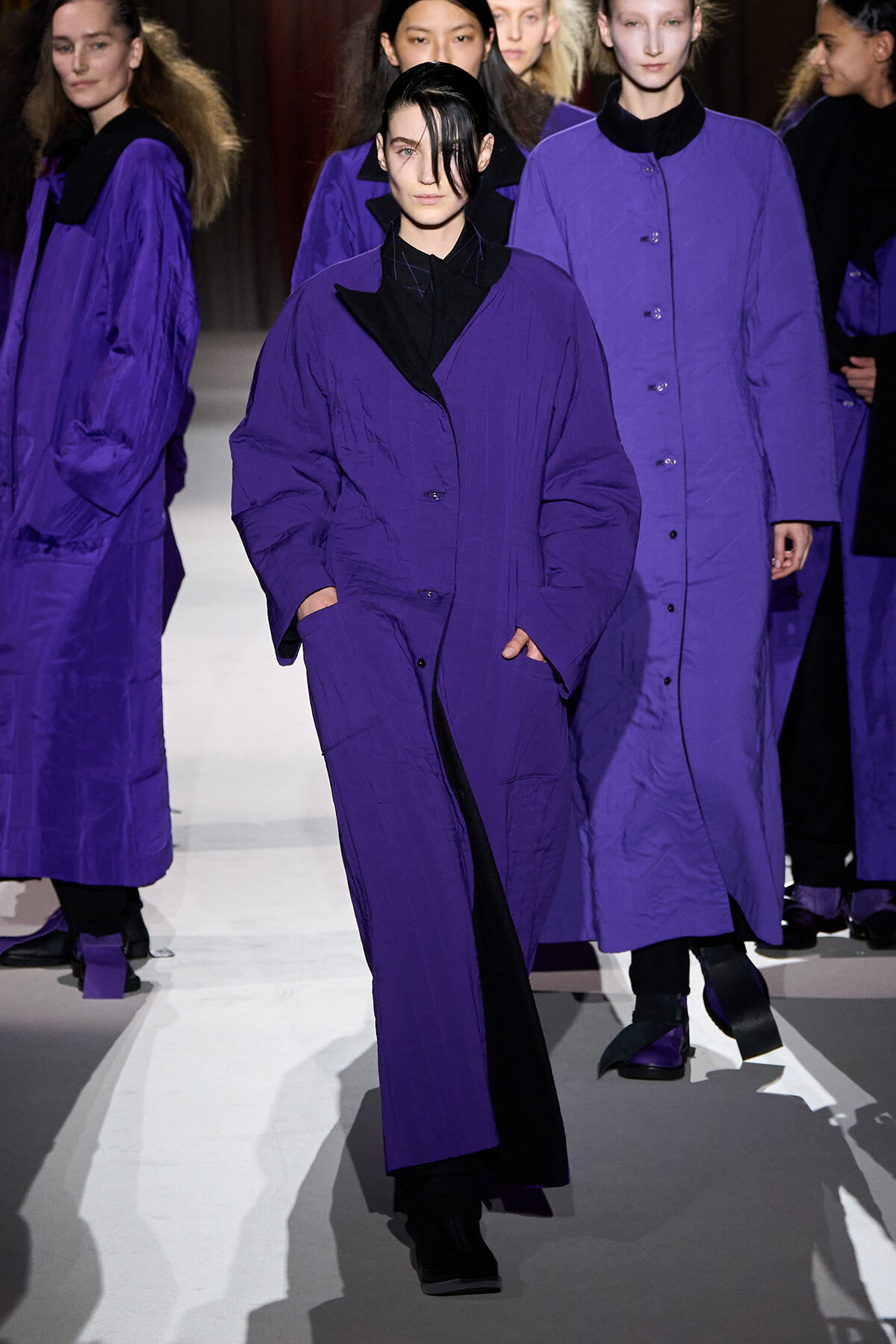 Models in matching long purple coats walk the runway, black outfits visible at the collar and cuffs.