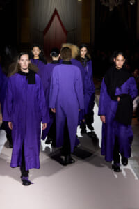 Models in matching purple coats walk down a runway during a fashion show, facing away from the camera.