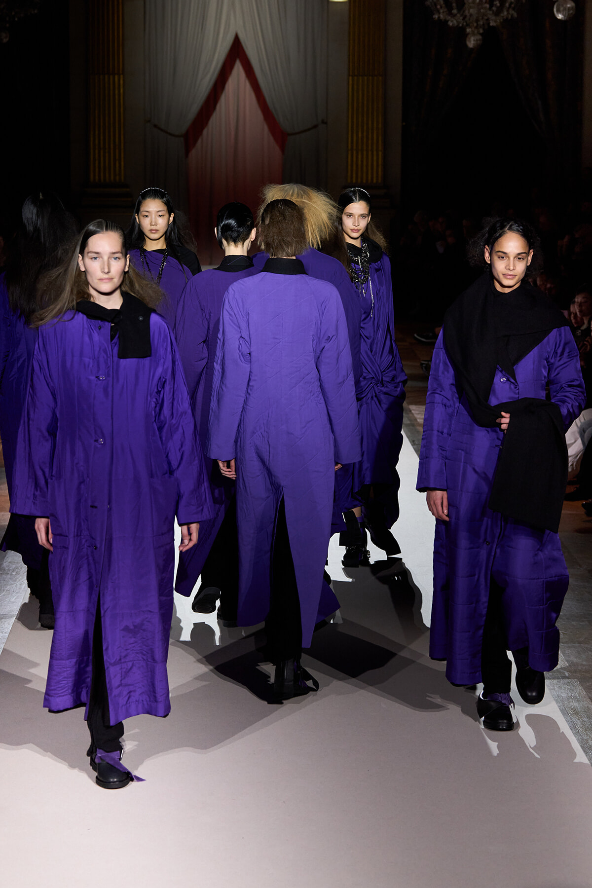 Models in matching purple coats walk down a runway during a fashion show, facing away from the camera.