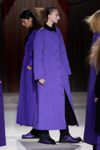Model wearing a long purple quilted coat on a fashion show runway.