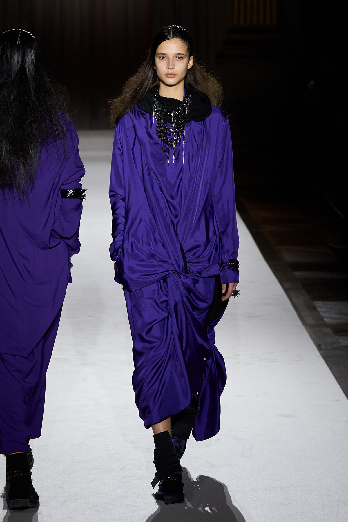 Model walking the runway in a loose, draped purple ensemble with a black scarf and layered black jewelry.