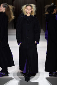 Model on a runway wearing a long black coat with purple lining showing at the cuffs and hem.