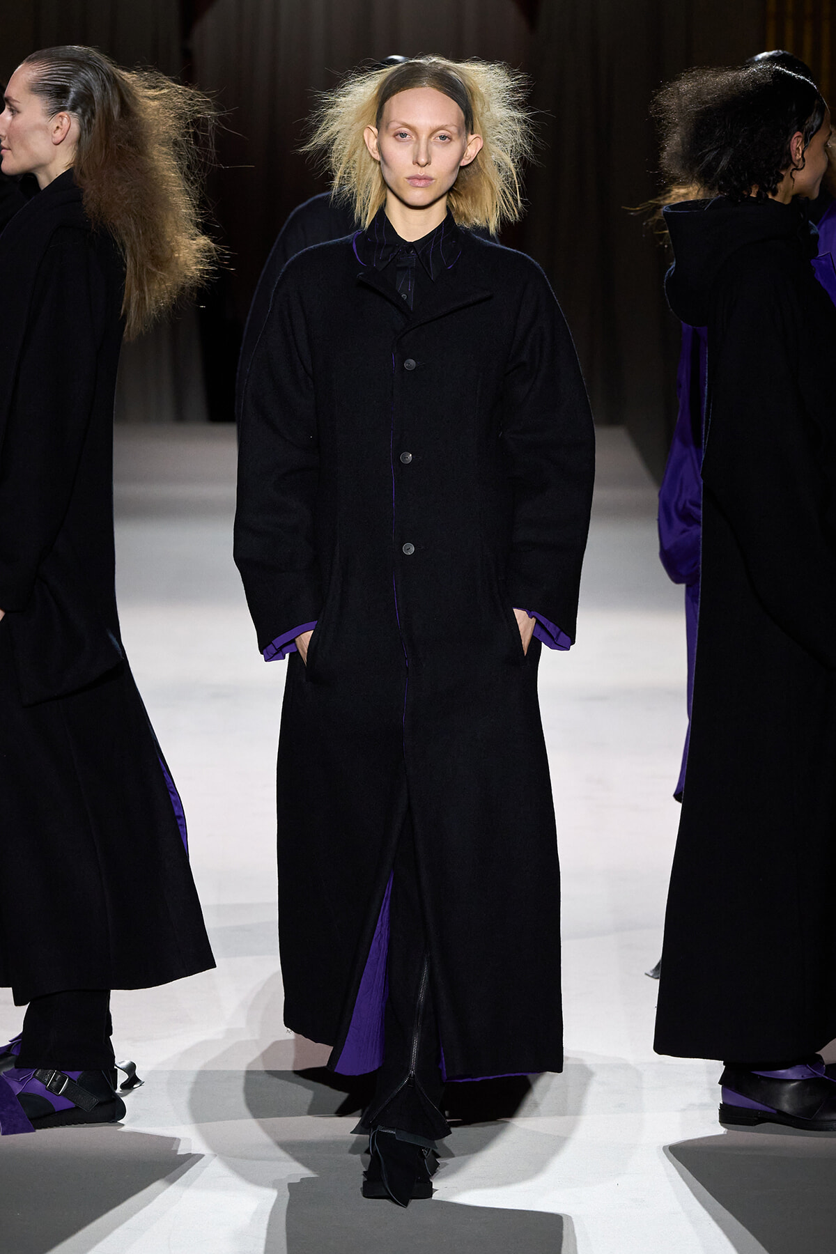 Model on a runway wearing a long black coat with purple lining showing at the cuffs and hem.