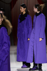 Women models wear matching long purple coats on a runway, black collars, purple shoes, and dark background shows a fashion show.