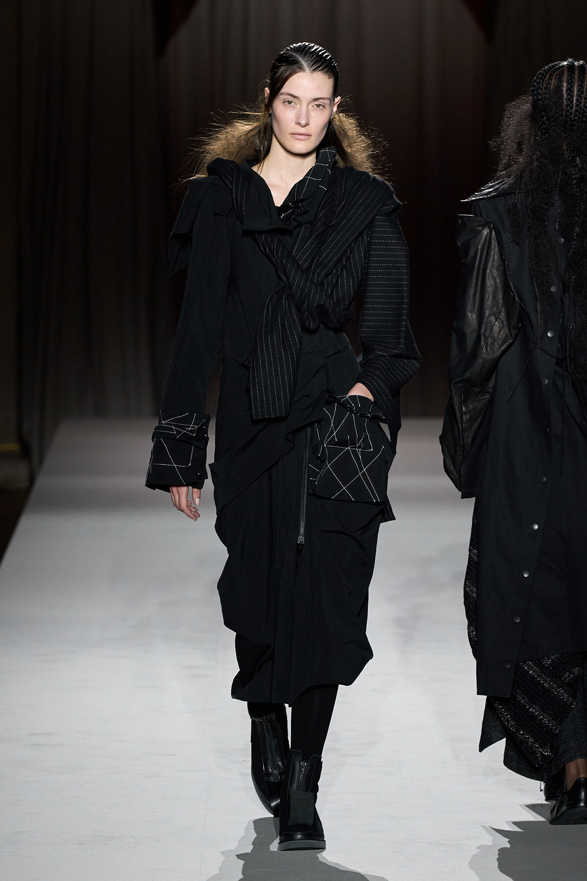 Model walking a runway in an all-black, layered outfit with oversized sleeves and boots.