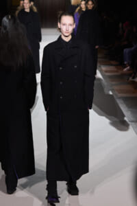 Model walks the runway wearing a long black double‑breasted coat with hands in pockets, spotlight on her pale face.