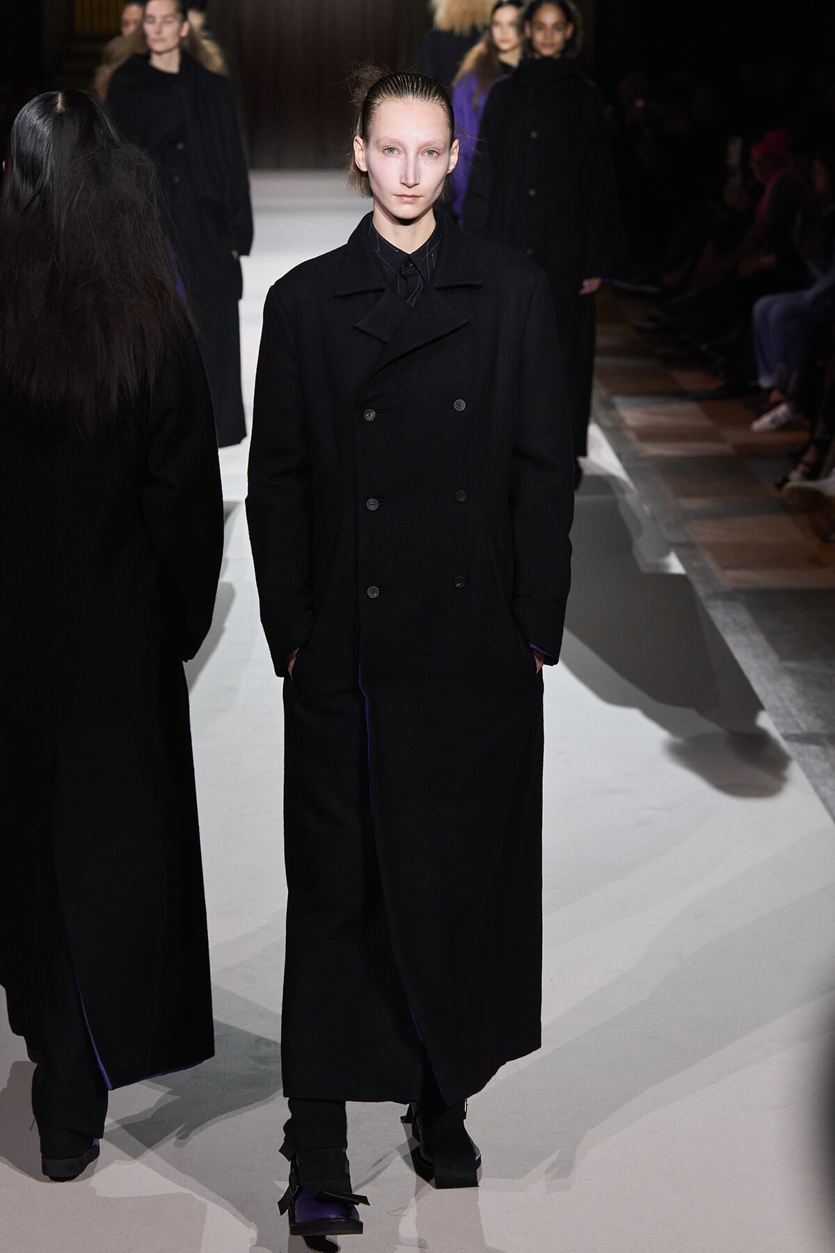 Model walks the runway wearing a long black double‑breasted coat with hands in pockets, spotlight on her pale face.