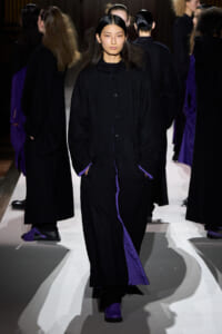 Female model walks a runway in a black button-front coat with purple lining visible at cuffs and hem, hands in pockets.