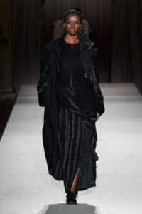 Model walks a runway in an all-black, multi-layered outfit: long coat over a zip-front top and a shimmering, draped skirt.