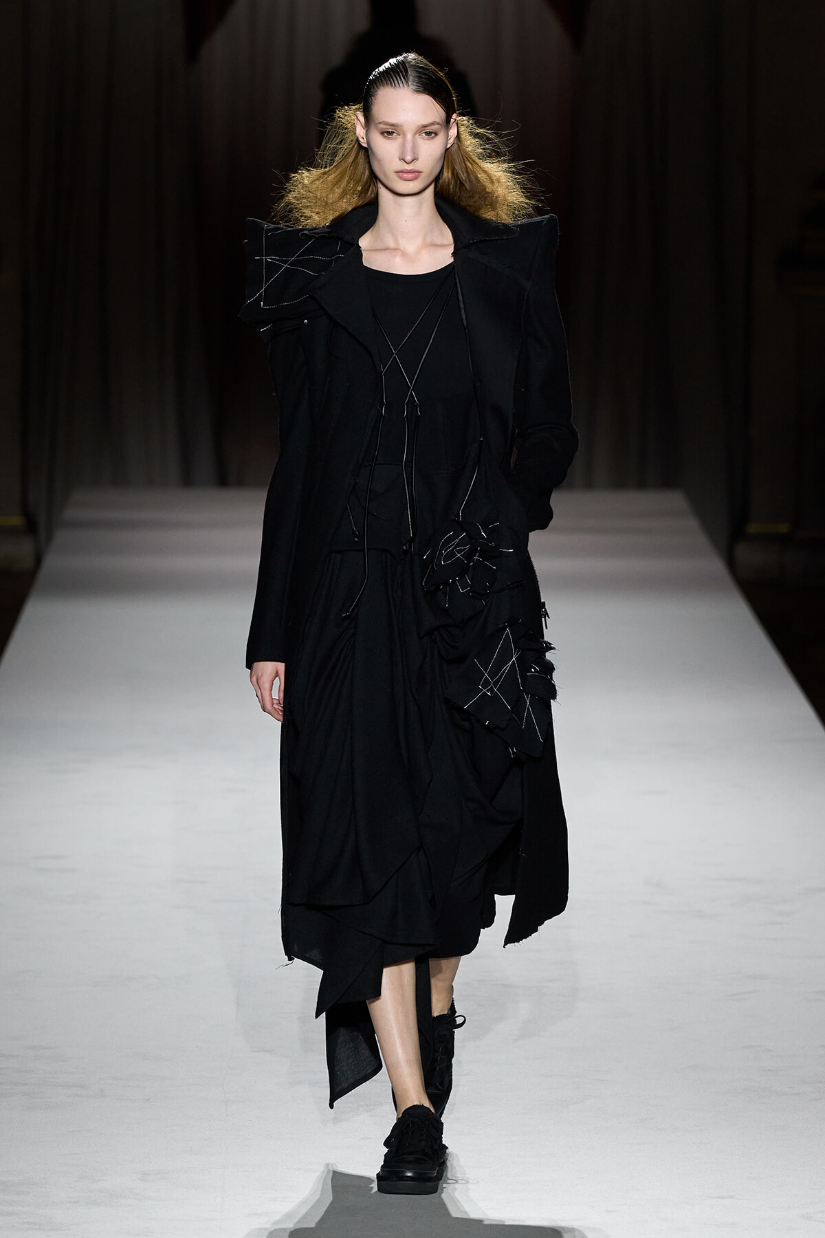 Model on a runway wearing an all-black, oversized coat with angular shoulder details and stitched panels, black shoes on a white catwalk.