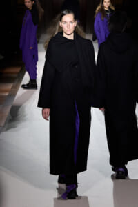 Model walks the runway in a long black coat with an oversized collar and purple accents visible underneath; other models in black outfits are in the background, on a white catwalk.