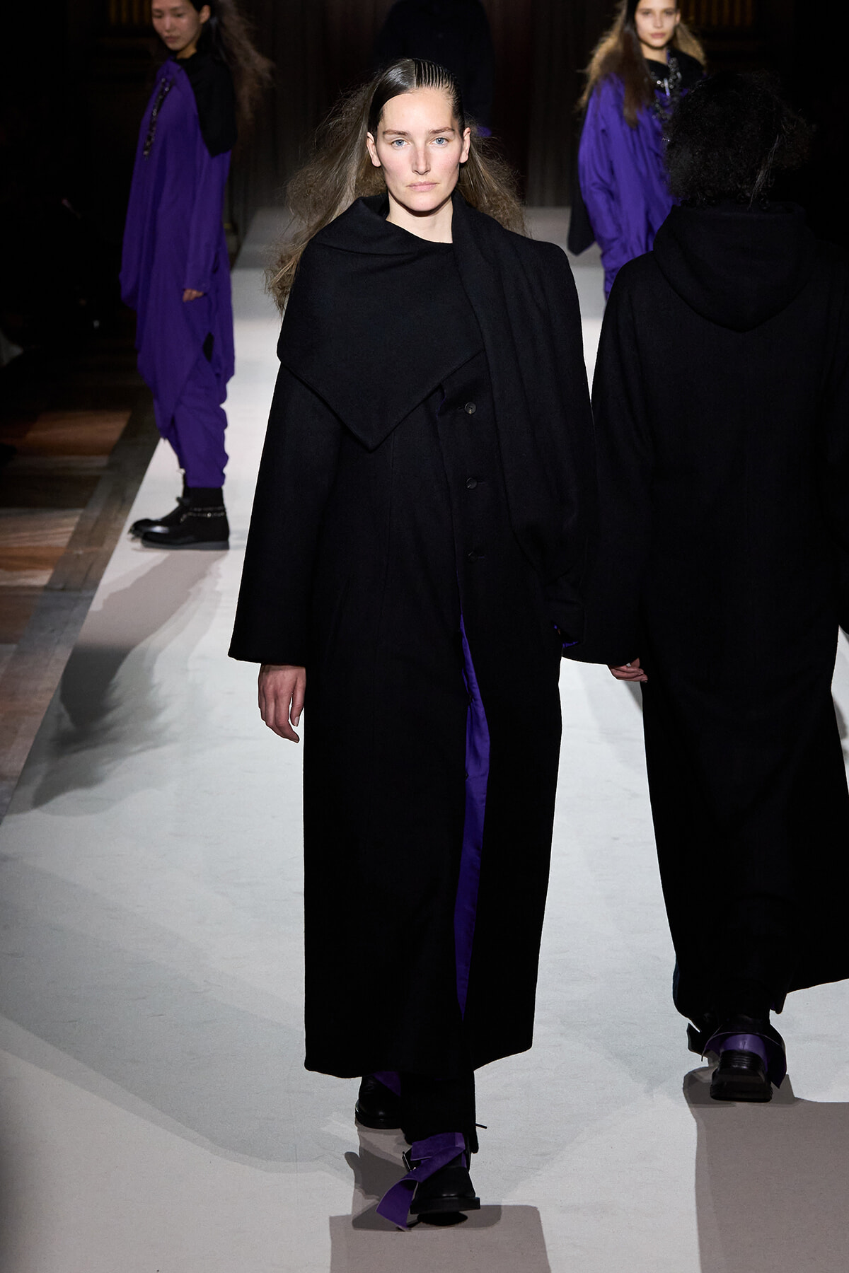 Model walks the runway in a long black coat with an oversized collar and purple accents visible underneath; other models in black outfits are in the background, on a white catwalk.