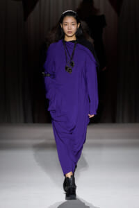Model walking a fashion runway in a loose, vibrant purple dress with layered black beaded necklaces and black boots.