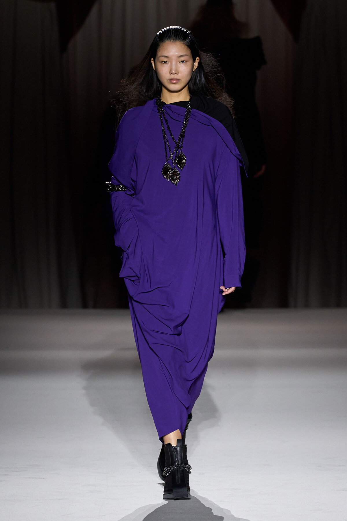 Model walking a fashion runway in a loose, vibrant purple dress with layered black beaded necklaces and black boots.