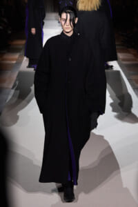 Model walking a runway in an oversized black coat with purple lining, hands in pockets, dark hair swept forward.
