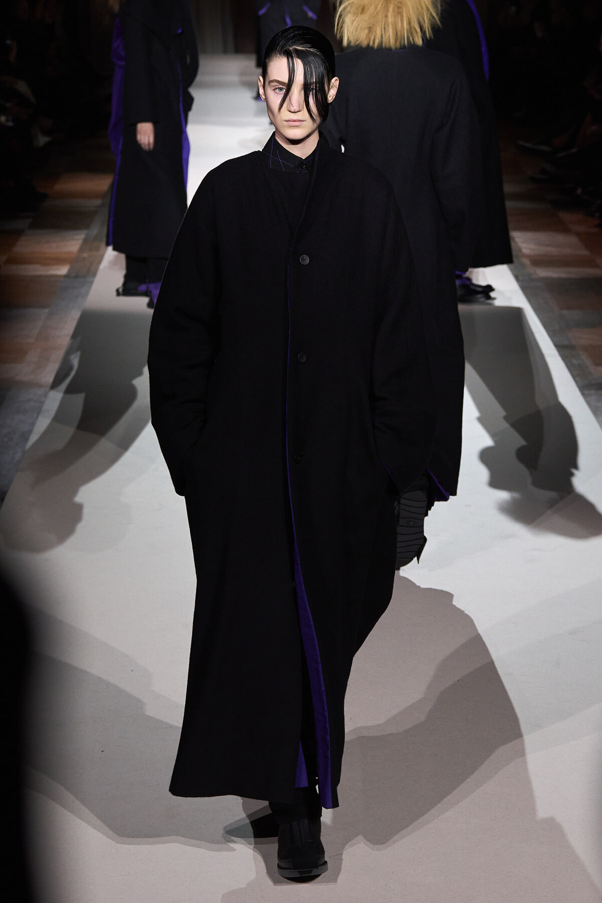 Model walking a runway in an oversized black coat with purple lining, hands in pockets, dark hair swept forward.
