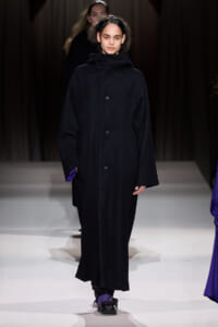 Model walking the runway in a long, oversized black coat with a high collar and subtle purple accents at the cuffs and lining.