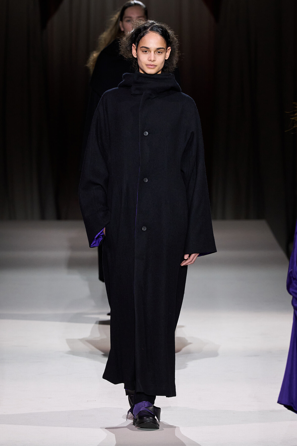 Model walking the runway in a long, oversized black coat with a high collar and subtle purple accents at the cuffs and lining.