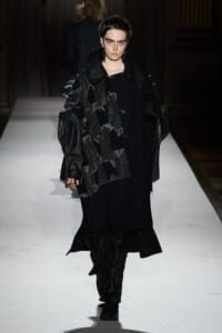 Model walking a runway in an all-black ensemble with a textured scarf and oversized leather-trimmed coat.