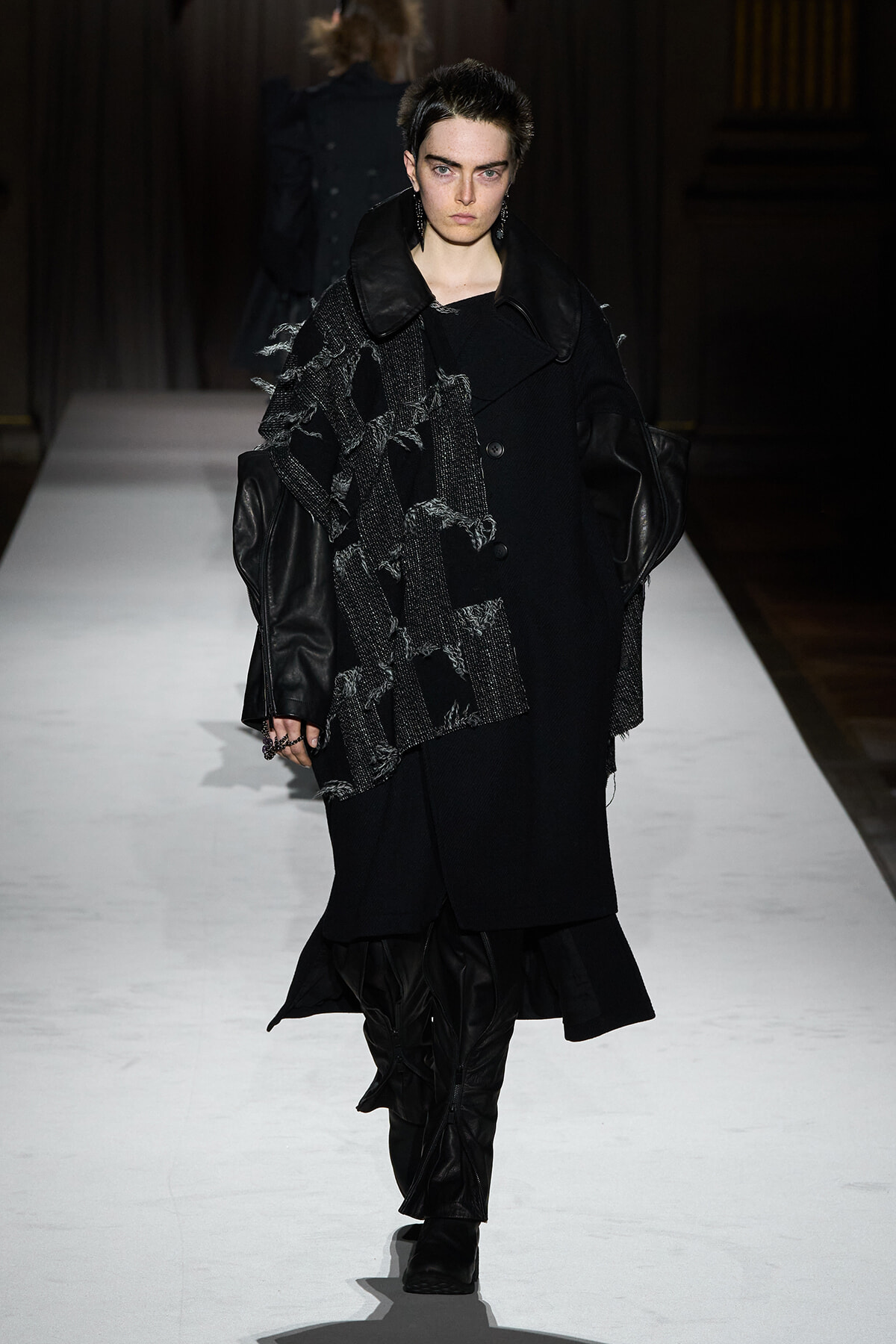 Model walking a runway in an all-black ensemble with a textured scarf and oversized leather-trimmed coat.