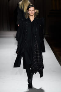 Model walking runway in an oversized black coat with dramatic shoulders and a plaid skirt peeking from underneath, dark minimalist styling.
