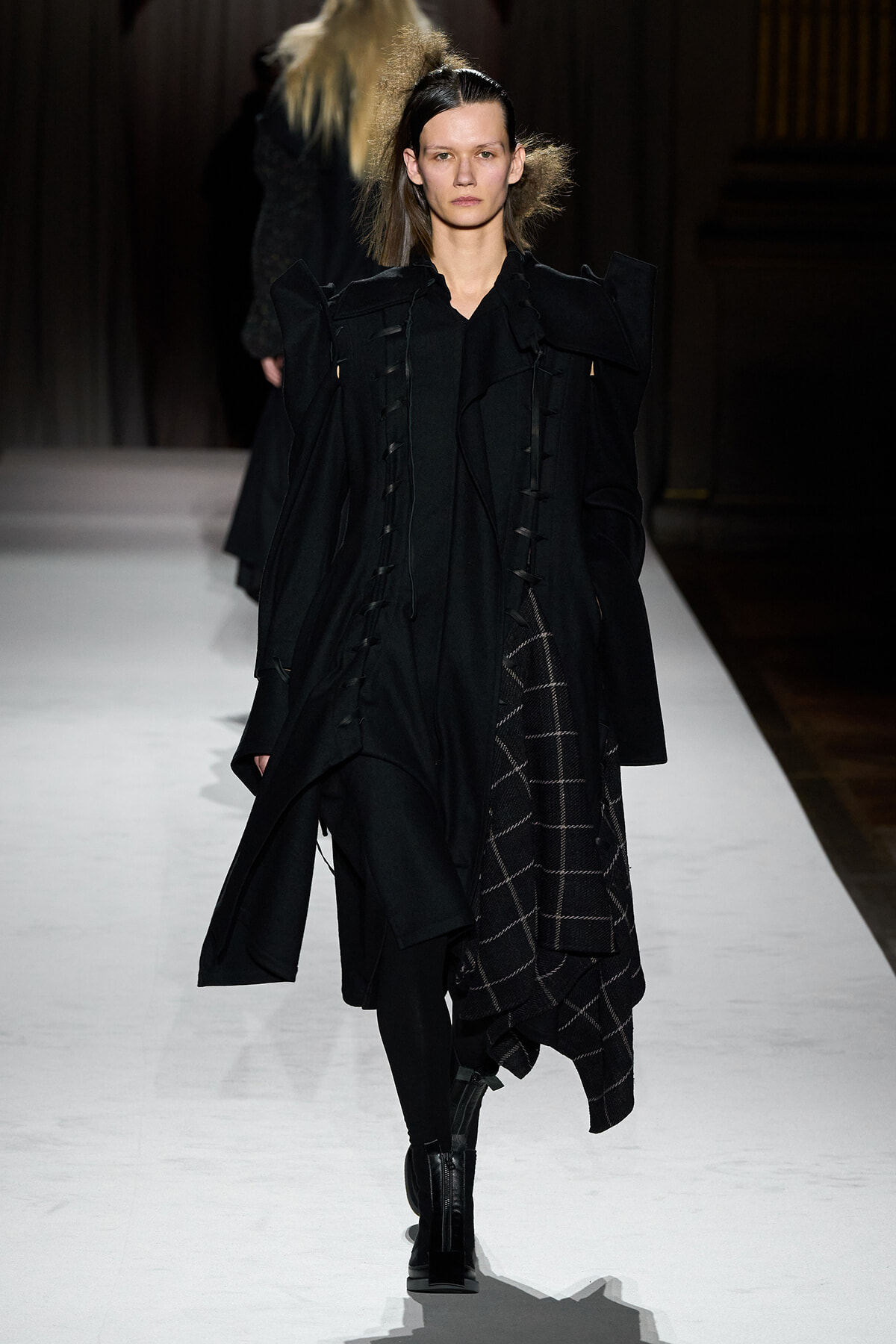 Model walking runway in an oversized black coat with dramatic shoulders and a plaid skirt peeking from underneath, dark minimalist styling.