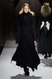 Female model walks a runway in a long black layered coat with ruffles and glittery sleeves.