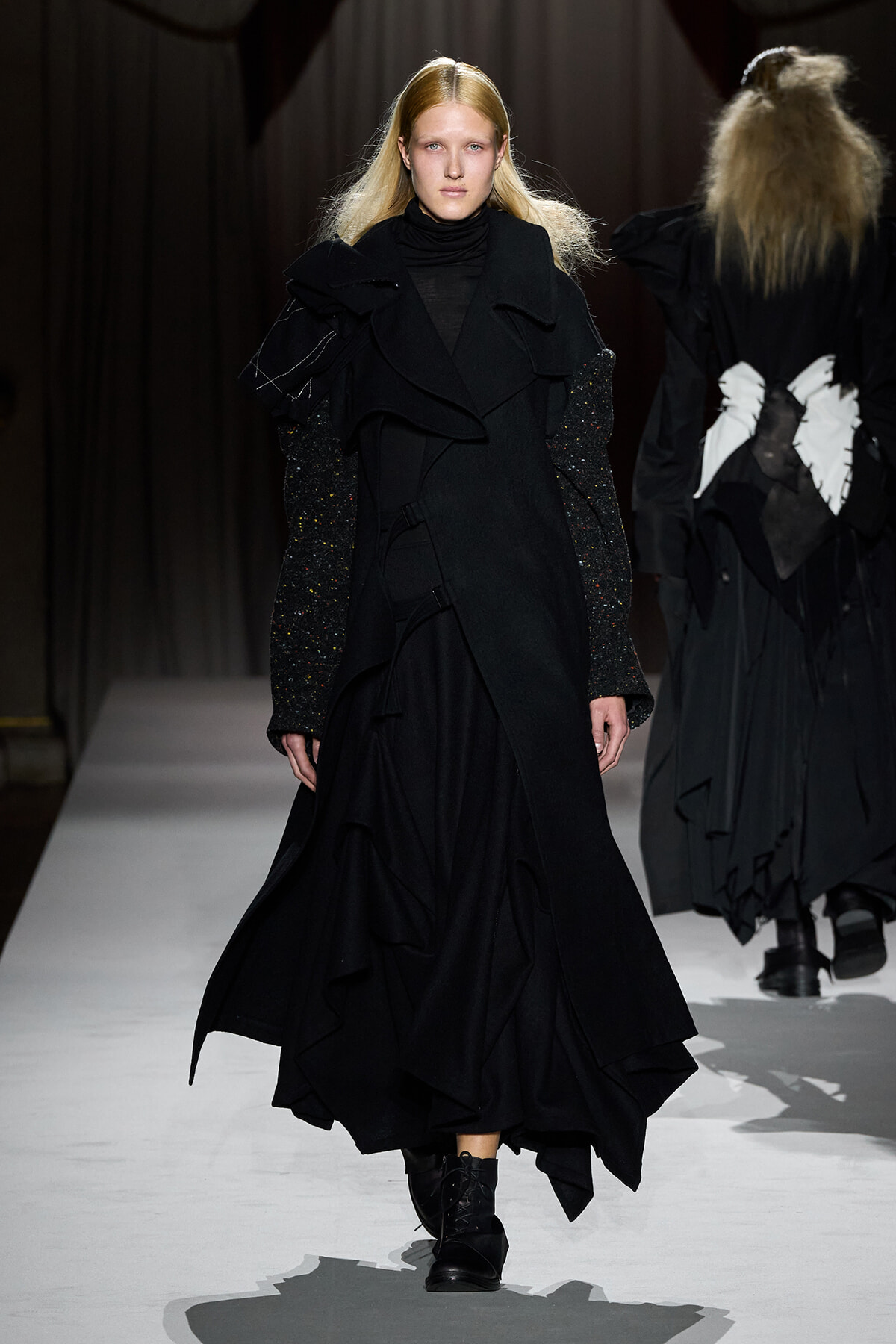 Female model walks a runway in a long black layered coat with ruffles and glittery sleeves.