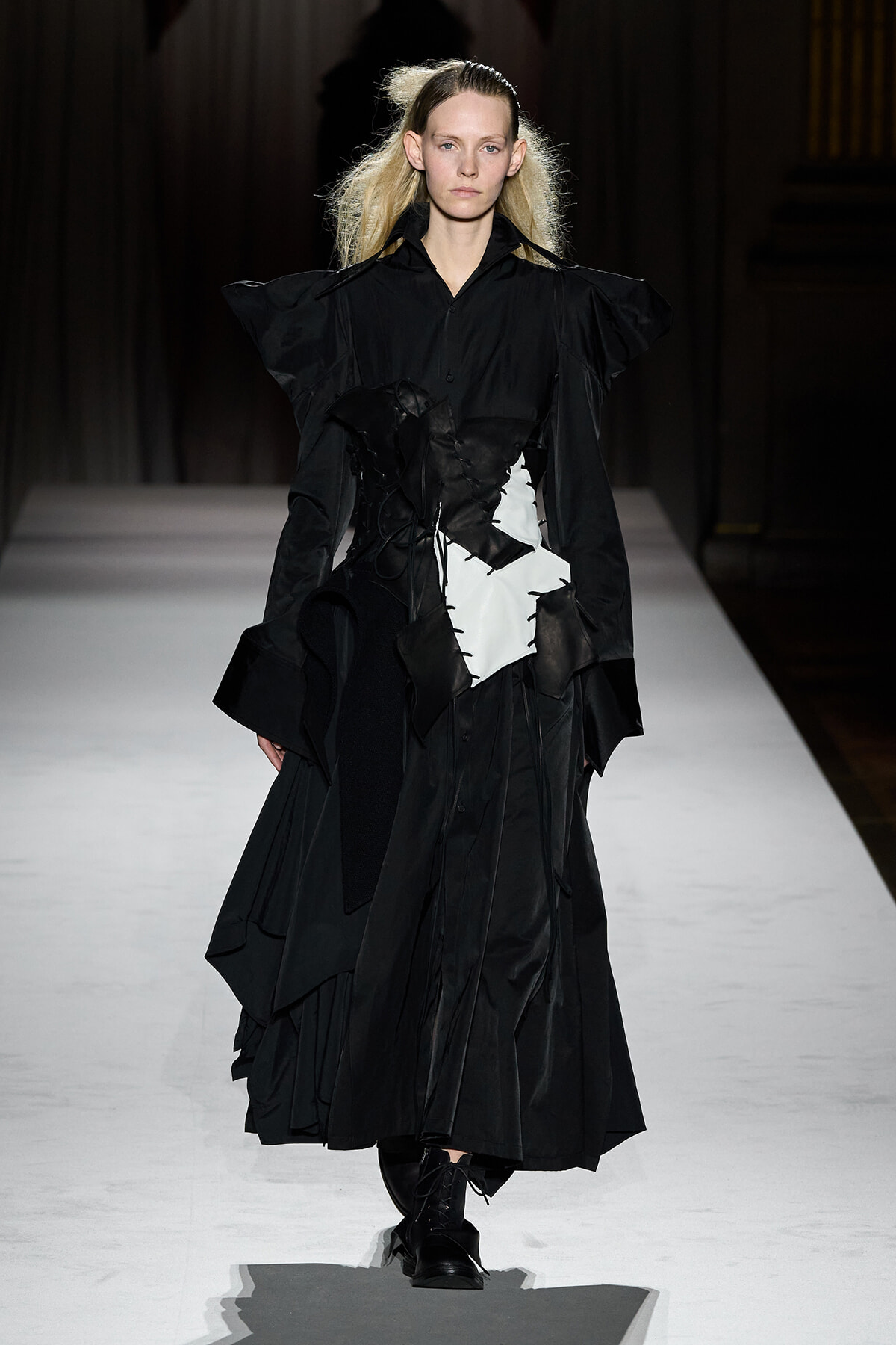 Model walking the runway in a dramatic black avant-garde gown with structured shoulders and a white abstract panel, dark background.