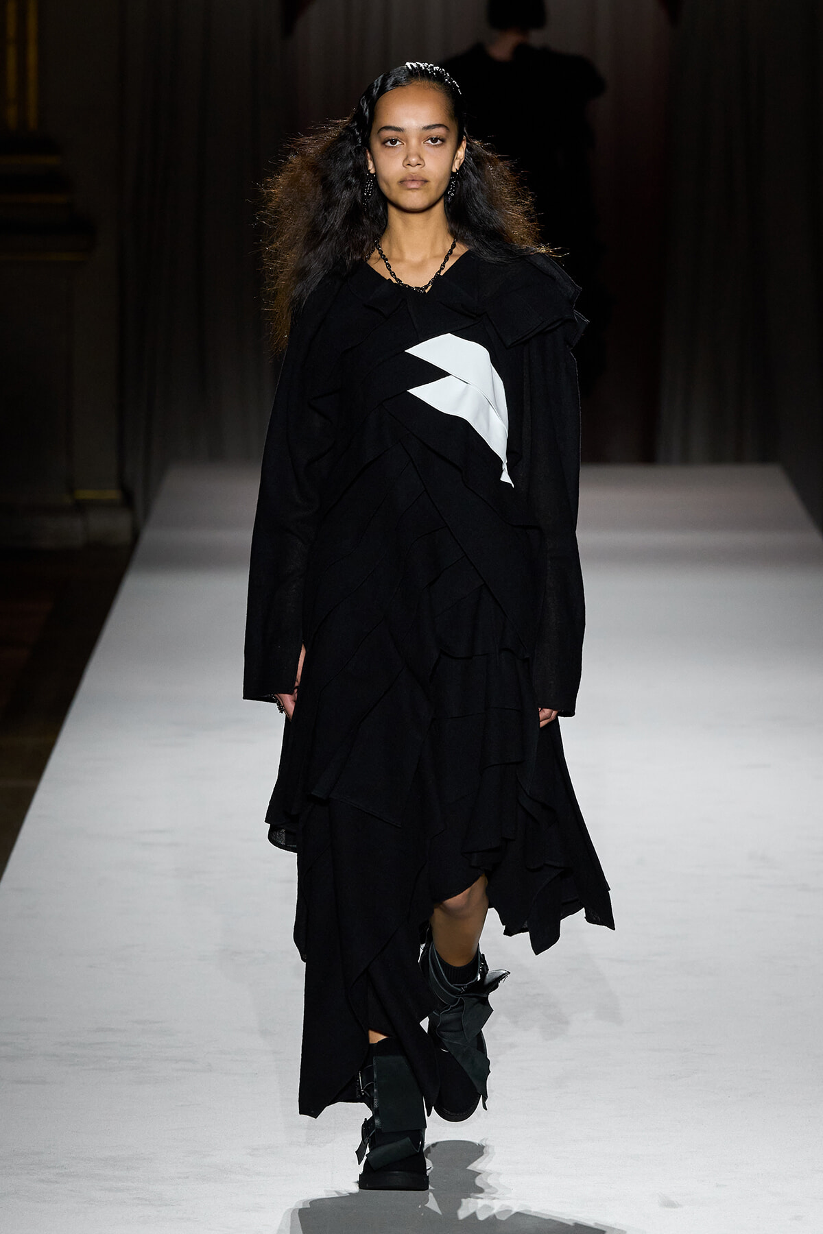 Model walking a fashion runway in a black, layered gown with a white chevron detail and chunky boots.