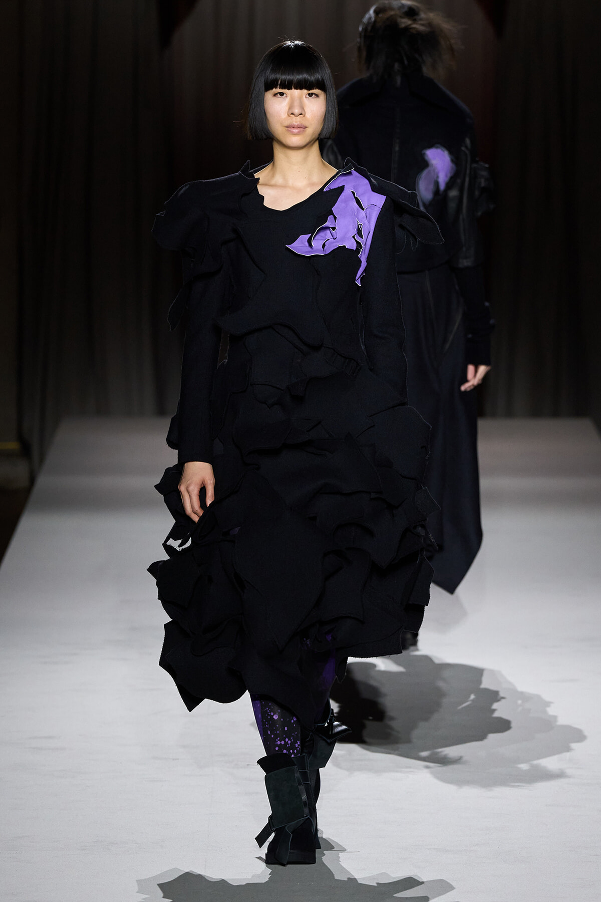 Model walking a fashion runway in a black ruffled gown with purple appliqué on the shoulder/torso area. edgy, avant-garde silhouette.