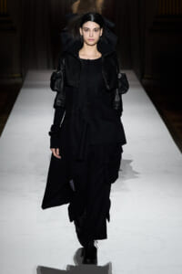 Model walks a fashion runway in an all-black, layered ensemble with oversized sleeves and an asymmetric, draped skirt.