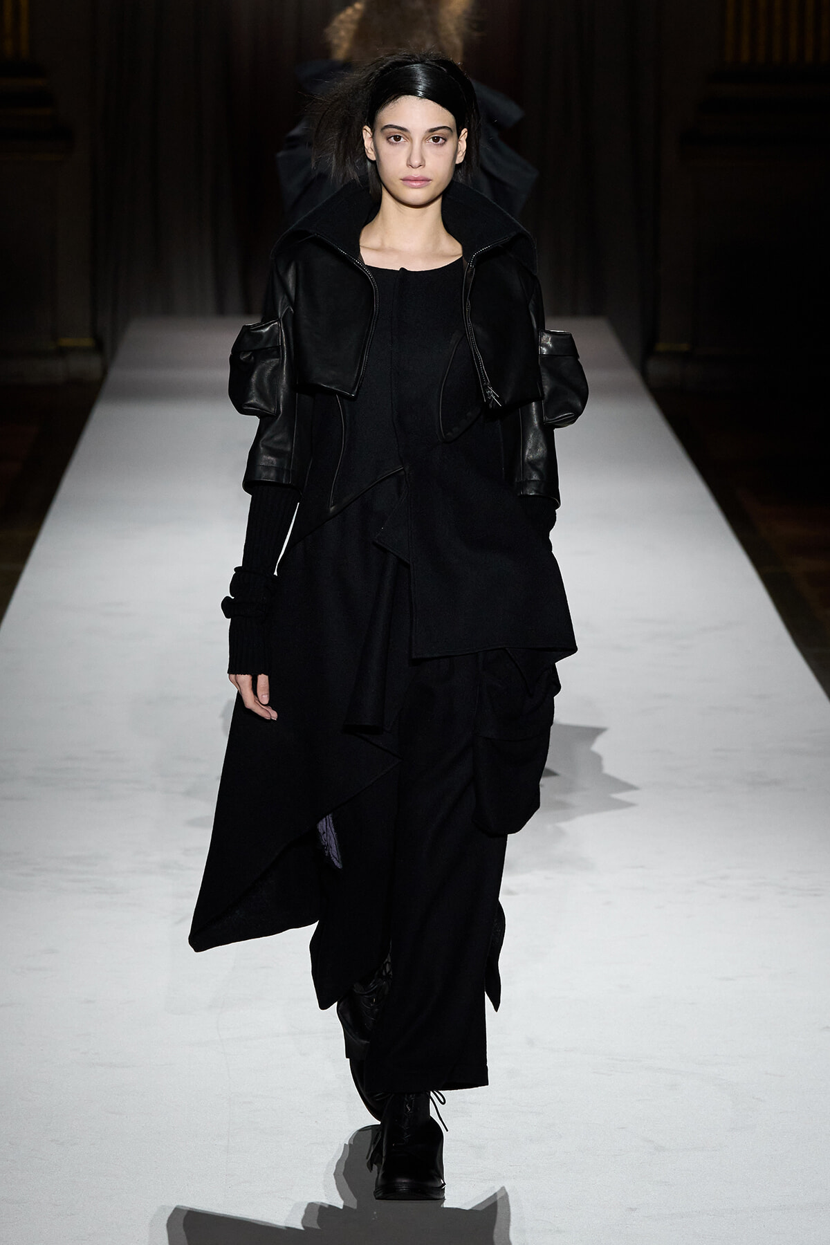 Model walks a fashion runway in an all-black, layered ensemble with oversized sleeves and an asymmetric, draped skirt.