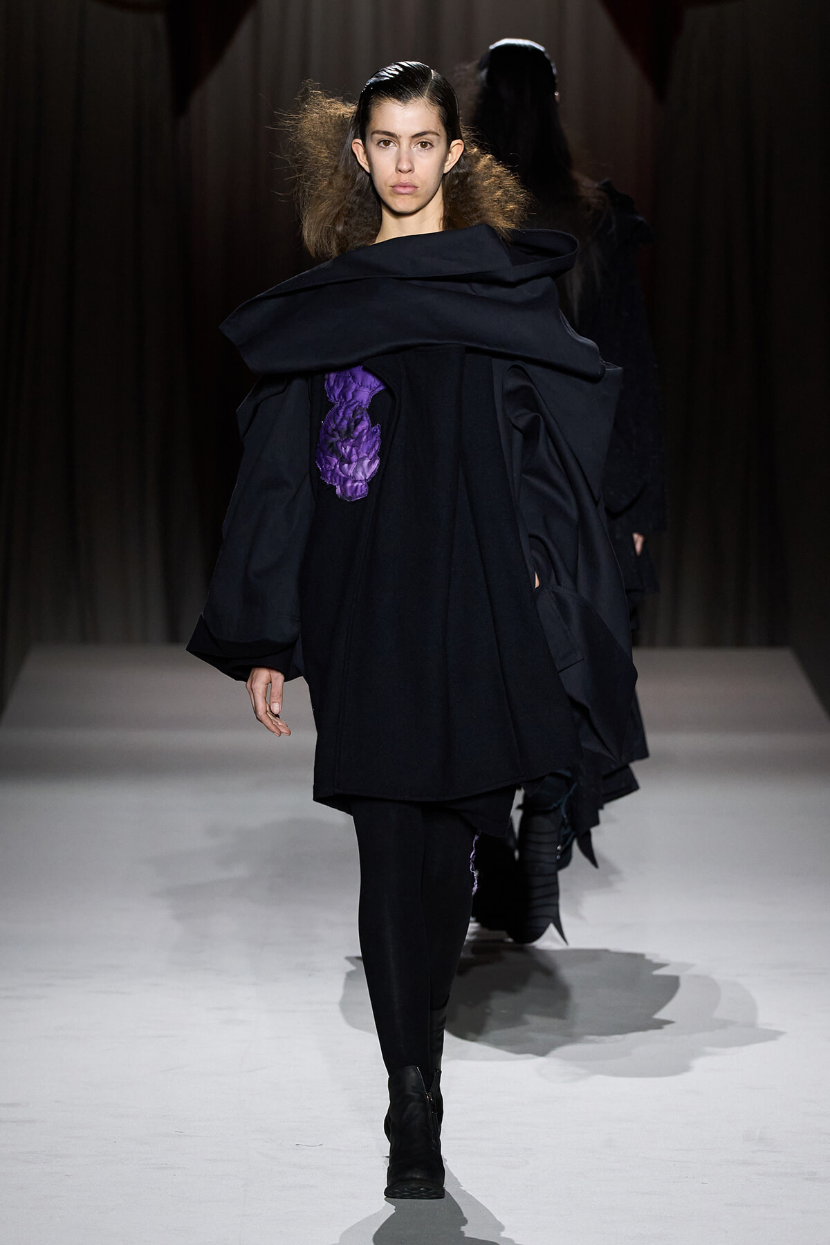 Model walking a runway in a black oversized coat with a purple stitched accent on the left chest area.