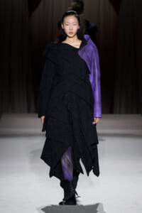 Model on a runway wearing a black, layered, asymmetrical dress with a single purple sleeve and black ankle boots.