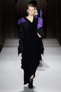 Model walks a runway in a black ruffled gown with purple angular shoulder accents.