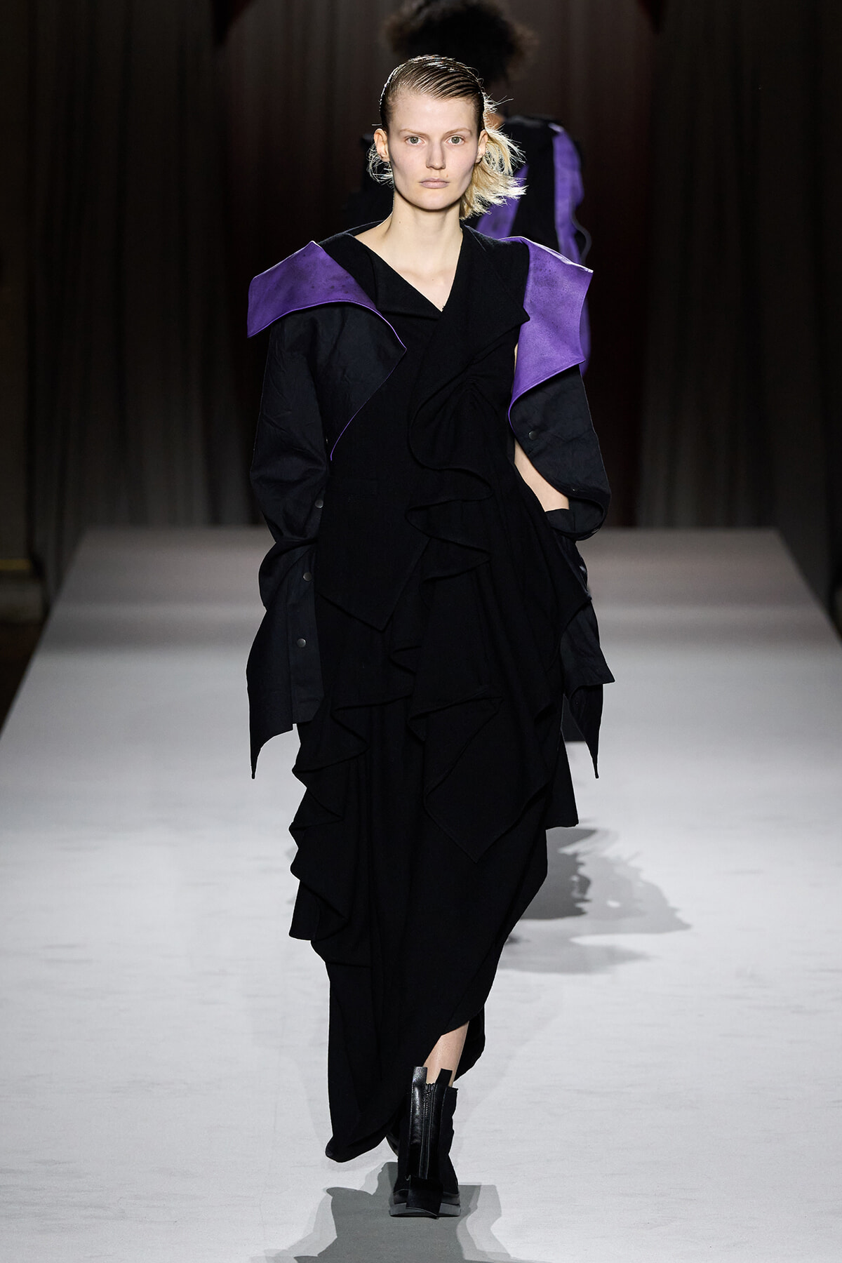 Model walks a runway in a black ruffled gown with purple angular shoulder accents.