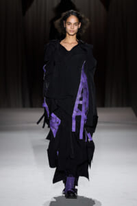 Model walks the runway in a black layered avant-garde coat with purple ribbon accents.