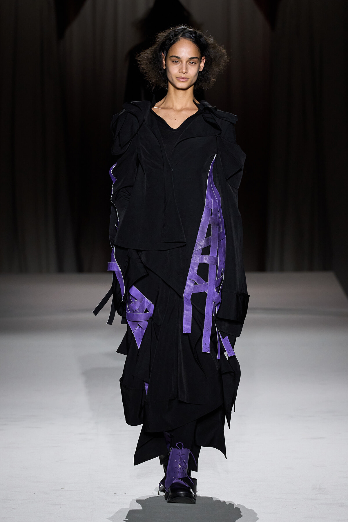 Model walks the runway in a black layered avant-garde coat with purple ribbon accents.