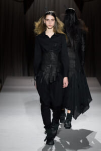 Front-model walks the runway in an all-black avant-garde outfit with a corset-like bodice and layered hem.