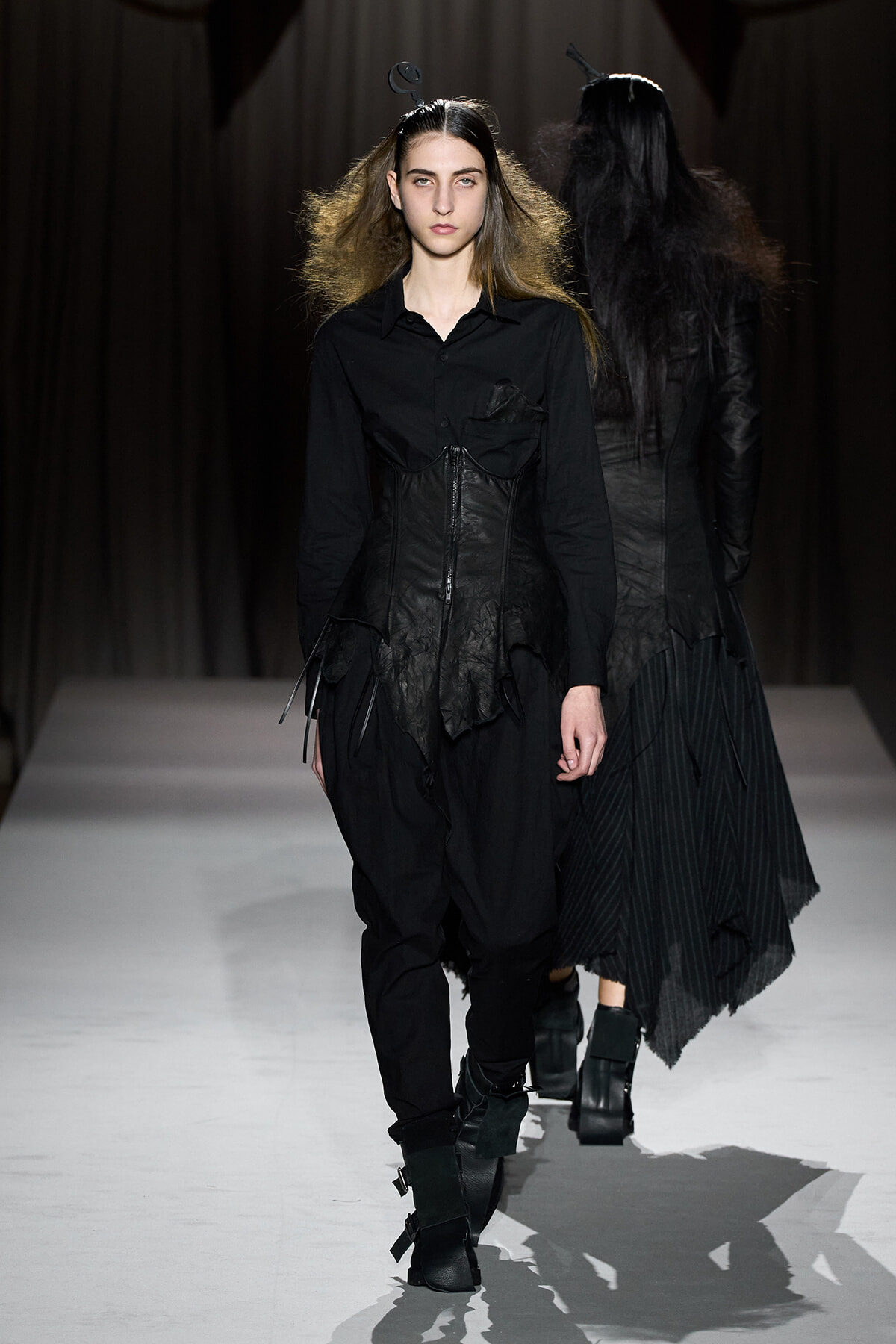 Front-model walks the runway in an all-black avant-garde outfit with a corset-like bodice and layered hem.