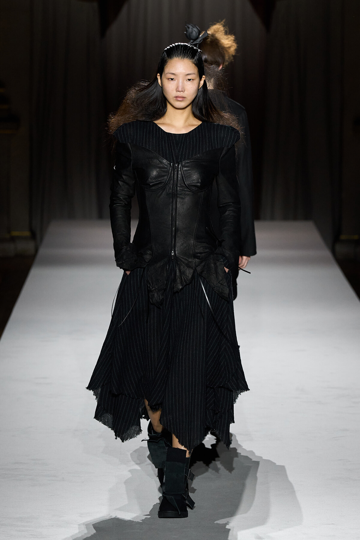 Female model walking a fashion runway in a black sculpted leather corset and layered pinstripe skirt with tall boots.