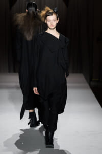 Model walks a fashion runway in an all-black, oversized outfit with a layered draped collar and black ankle boots.