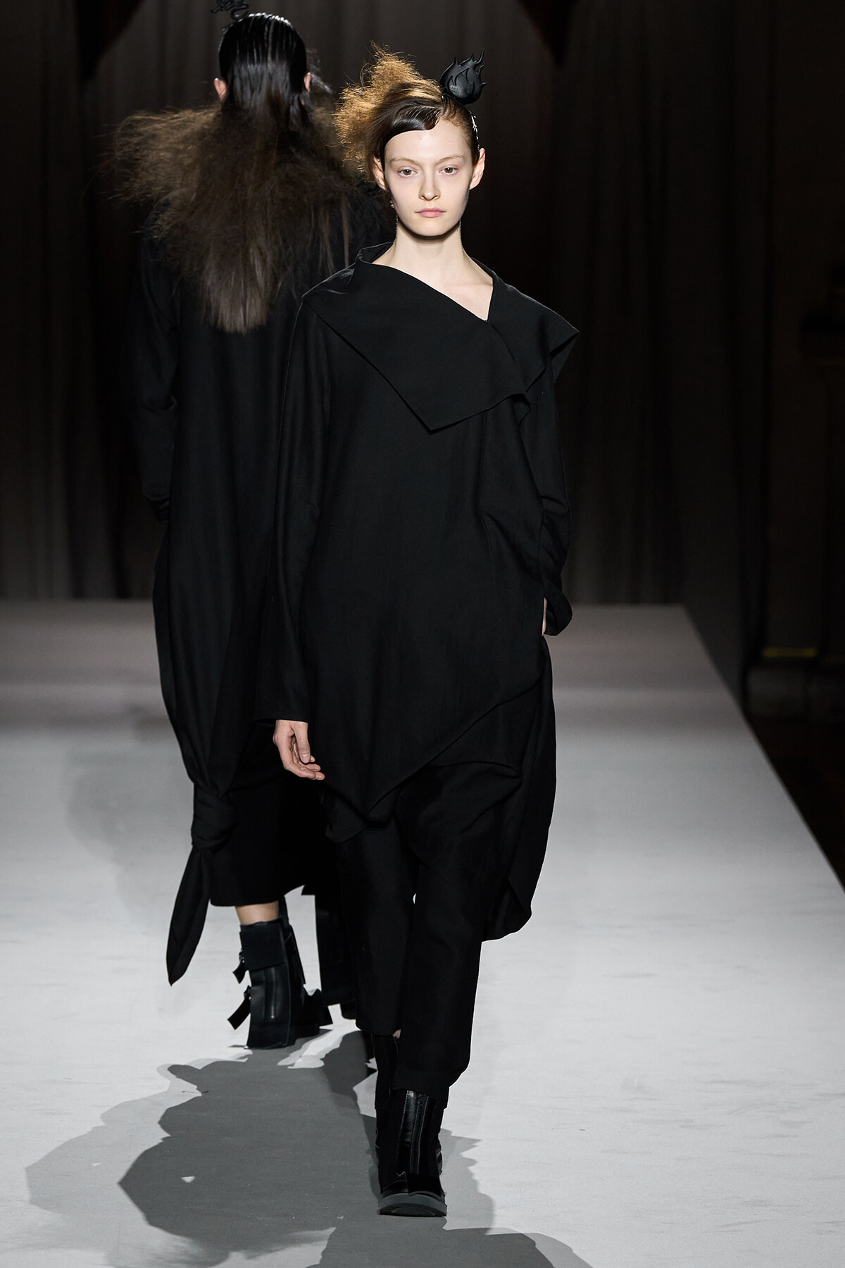 Model walks a fashion runway in an all-black, oversized outfit with a layered draped collar and black ankle boots.