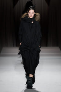 Model walking the runway in an all-black, draped outfit with a front knot and oversized scarf.