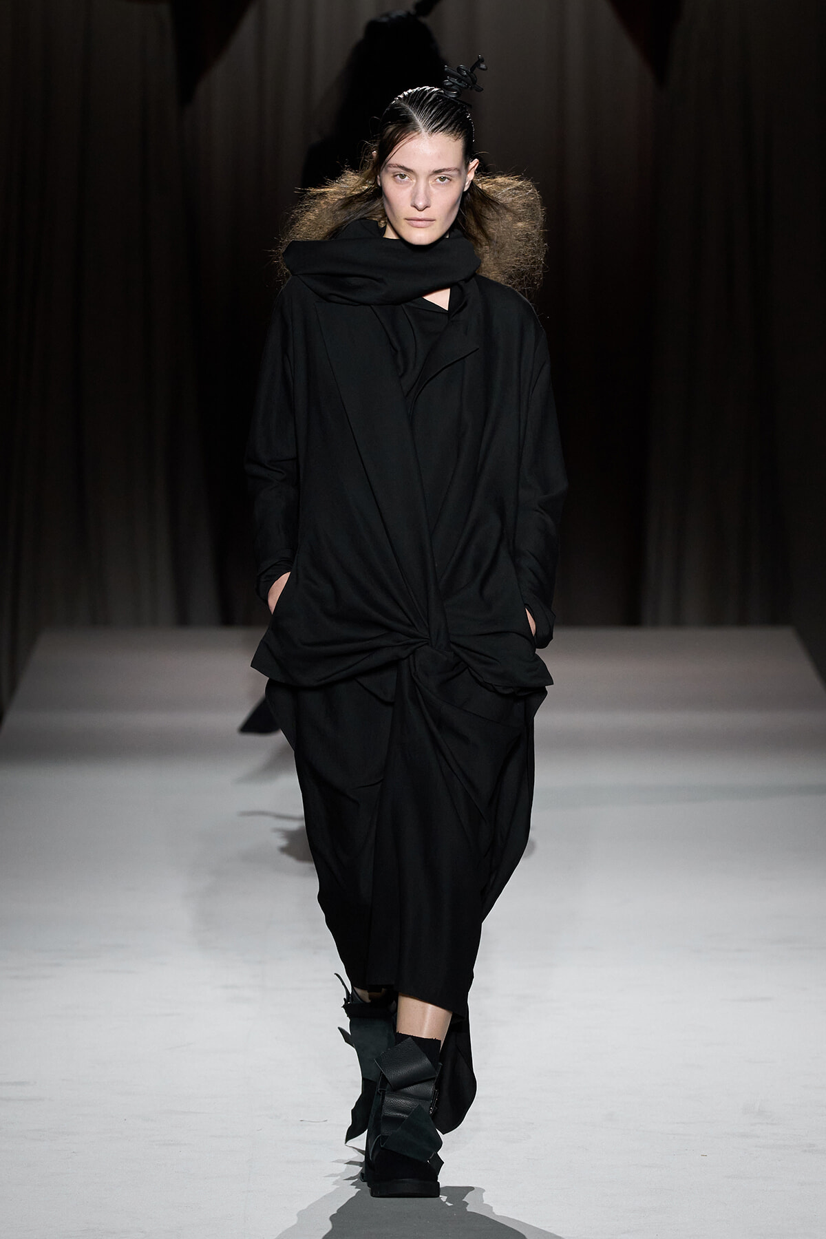 Model walking the runway in an all-black, draped outfit with a front knot and oversized scarf.