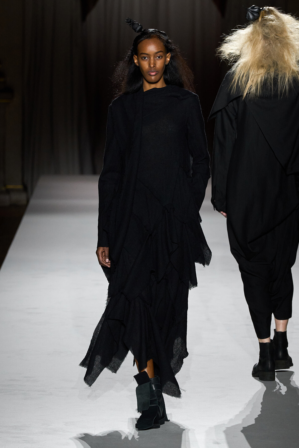 Female model walks the runway in a long, black layered dress with ankle boots on a white catwalk.