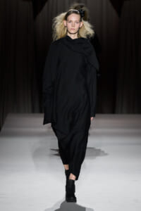 Model walking runway in oversized black draped outfit with black ankle boots, minimalist monochrome look.