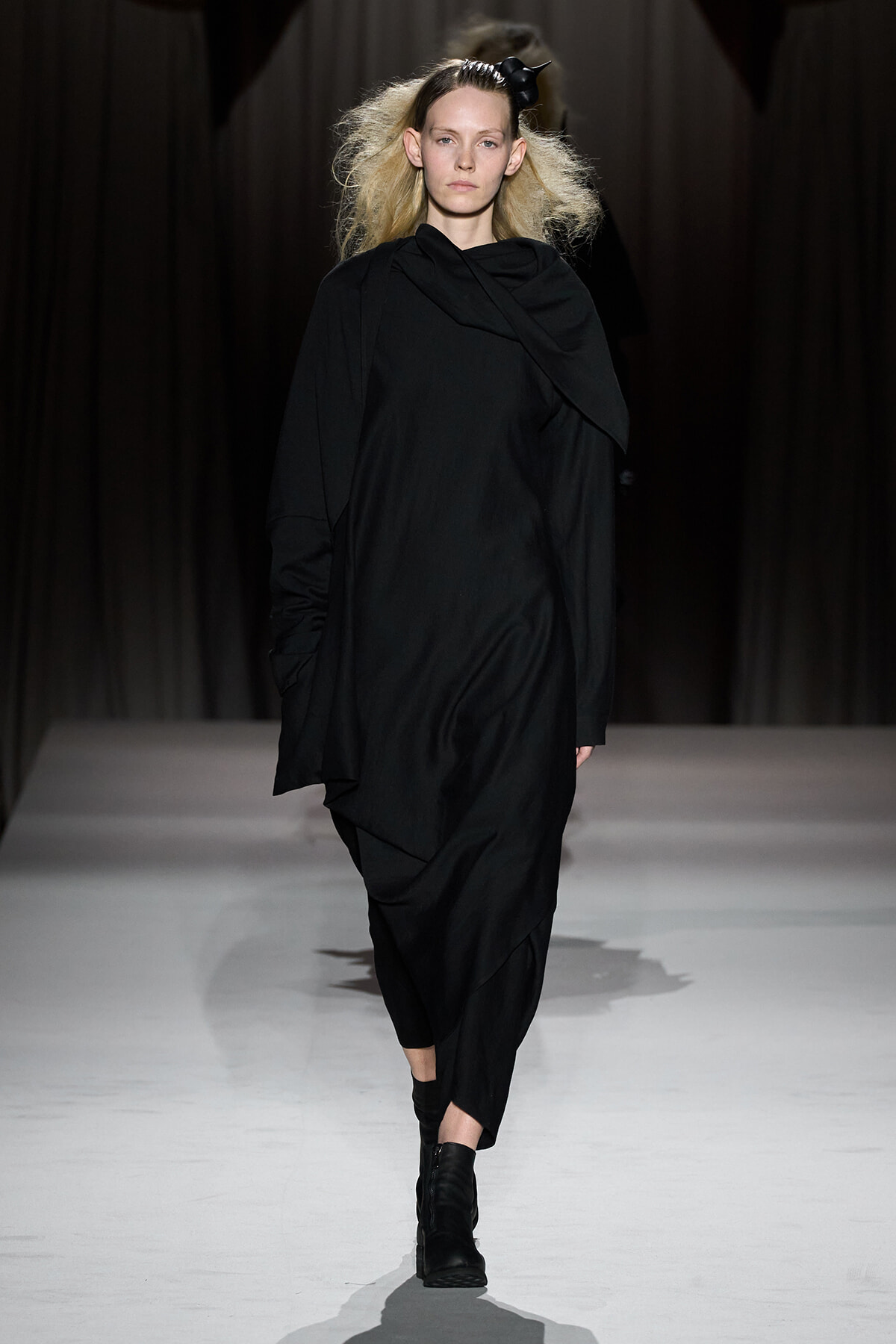 Model walking runway in oversized black draped outfit with black ankle boots, minimalist monochrome look.